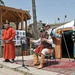 A traditional Mongolian cultural performance is taking place outdoors, likely at a military base or international event given the presence of soldiers in the background. Performers are dressed in traditional Mongolian attire including a singer in a red deel robe, a musician playing a morin khuur (horsehead fiddle), and two guards in historical Mongolian warrior armor holding spears and shields. A traditional ger (yurt) is visible in the background alongside a wooden gazebo.