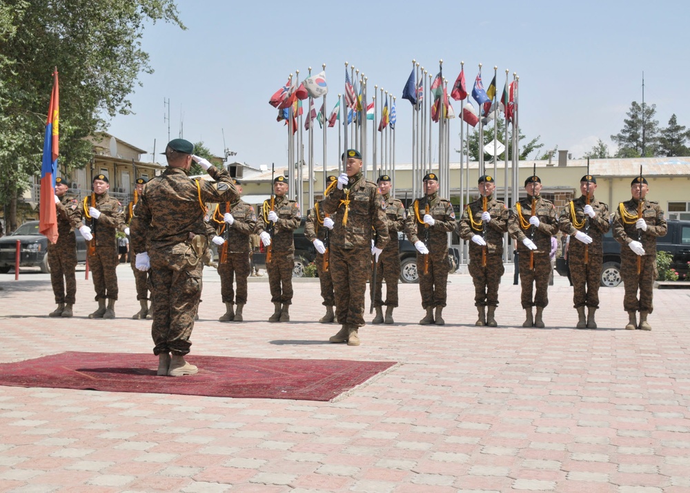 A military parade or formal ceremony is taking place in an outdoor courtyard, featuring soldiers in camouflage uniforms with green berets, white gloves, and yellow ceremonial lanyards. A commanding officer standing on a red carpet is saluting the formation of troops lined up before him. Multiple international flags are displayed on flagpoles in the background, suggesting a multinational military installation.