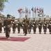 A military parade or formal ceremony is taking place in an outdoor courtyard, featuring soldiers in camouflage uniforms with green berets, white gloves, and yellow ceremonial lanyards. A commanding officer standing on a red carpet is saluting the formation of troops lined up before him. Multiple international flags are displayed on flagpoles in the background, suggesting a multinational military installation.
