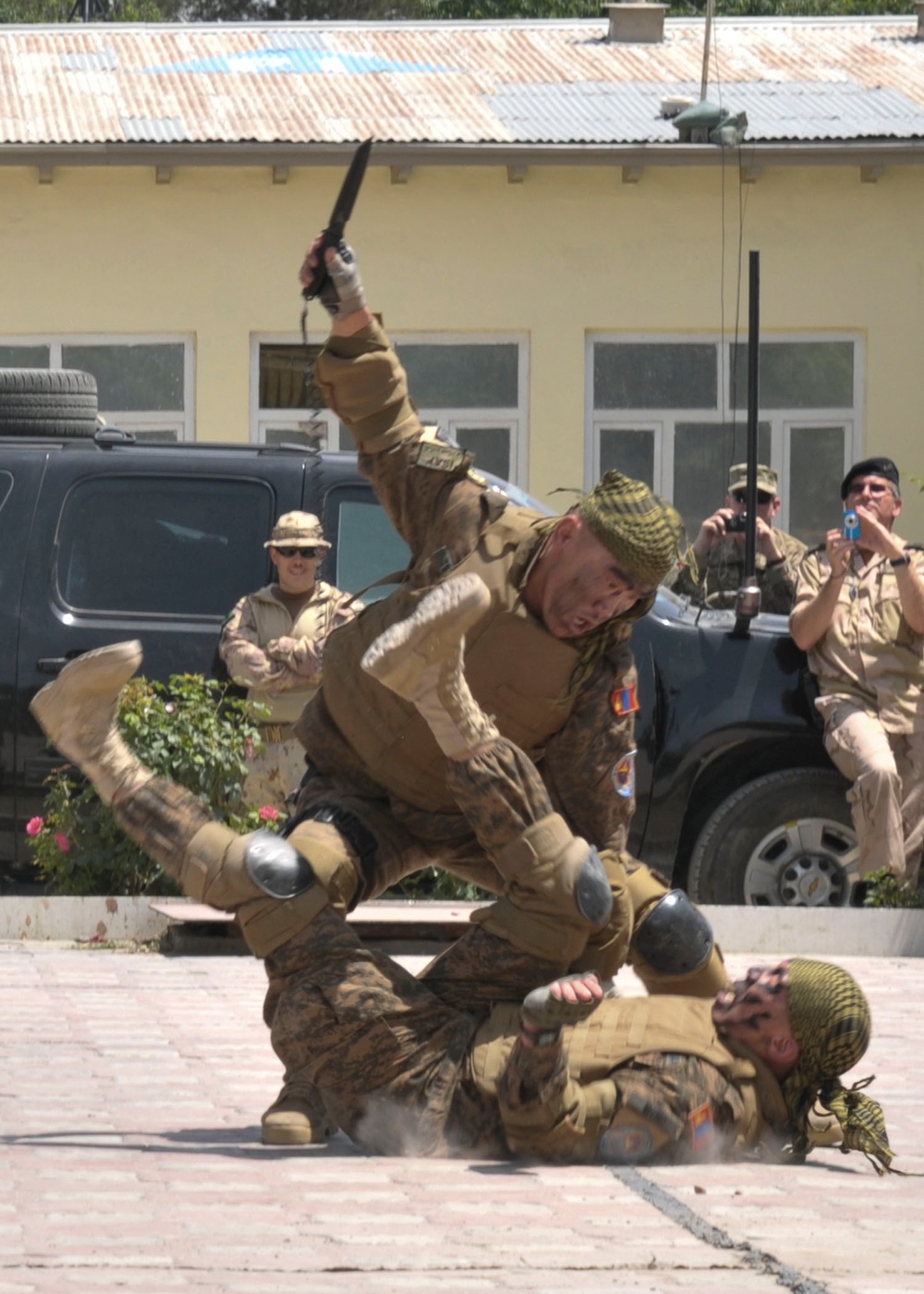 A military combat demonstration or training exercise is taking place on a paved outdoor area in front of a building. Two soldiers in camouflage uniforms are performing a knife combat drill, with one standing over the other who is on the ground. Several military observers in the background are photographing or watching the demonstration.