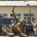 A military combat demonstration or training exercise is taking place on a paved outdoor area in front of a building. Two soldiers in camouflage uniforms are performing a knife combat drill, with one standing over the other who is on the ground. Several military observers in the background are photographing or watching the demonstration.
