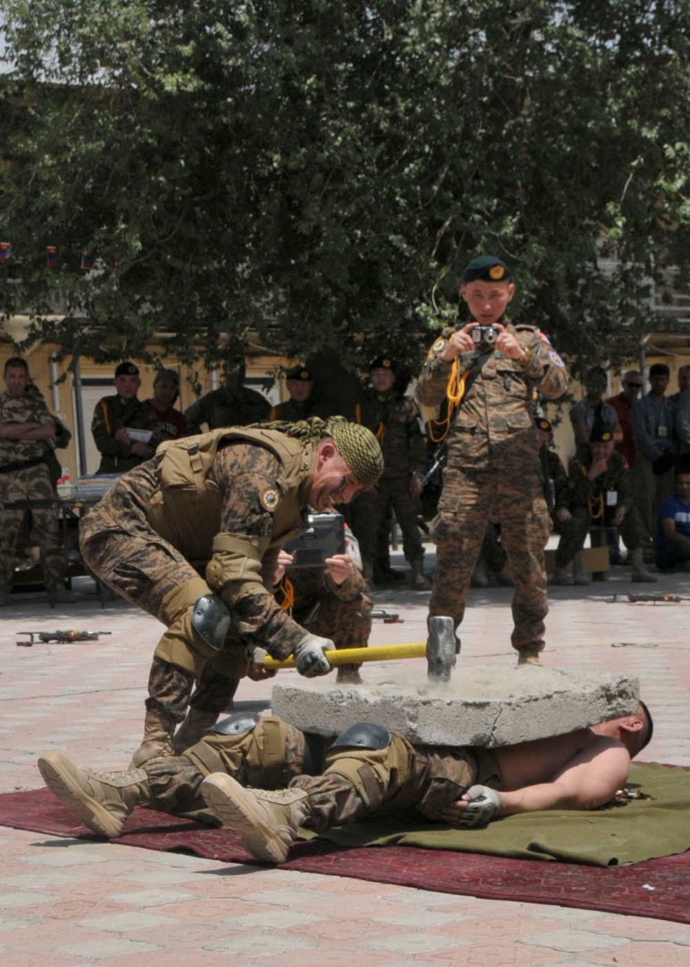 A military strength demonstration is taking place in an outdoor courtyard, where one uniformed soldier strikes a large concrete slab resting on a fellow soldier lying on a mat, using a sledgehammer. A crowd of uniformed military personnel watches from the background, and another soldier photographs the event. This appears to be a formal martial arts or combat skills demonstration for an assembled military audience.