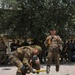 A military strength demonstration is taking place in an outdoor courtyard, where one uniformed soldier strikes a large concrete slab resting on a fellow soldier lying on a mat, using a sledgehammer. A crowd of uniformed military personnel watches from the background, and another soldier photographs the event. This appears to be a formal martial arts or combat skills demonstration for an assembled military audience.