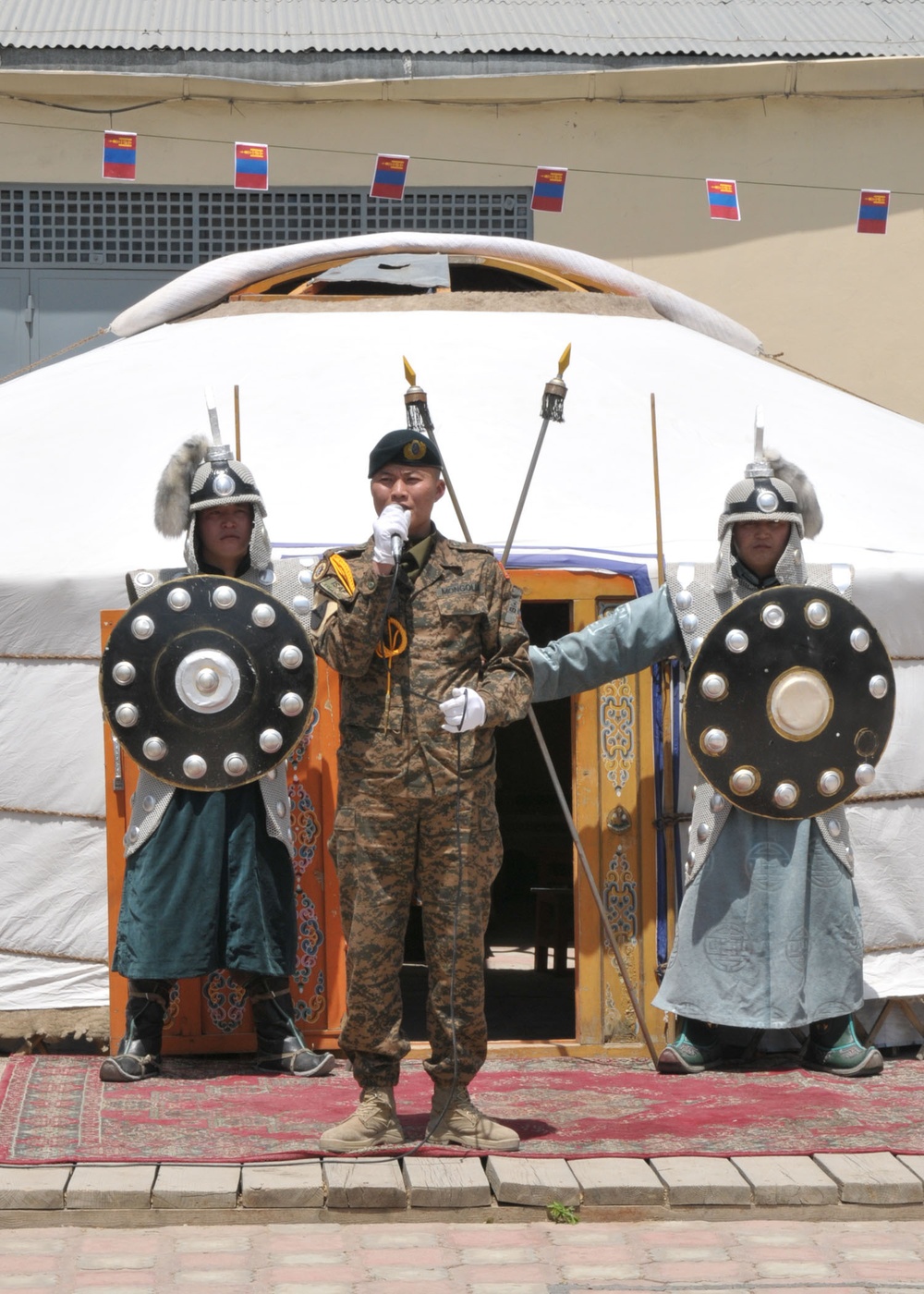 Three individuals stand in front of a traditional Mongolian ger (yurt) decorated with Mongolian flags on a string. The central figure wears a modern Mongolian military uniform and speaks into a microphone, while the two flanking figures wear traditional ceremonial armor including chainmail helmets and carry round decorated shields and spears. The setting appears to be a formal ceremonial or official event.