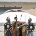 Three individuals stand in front of a traditional Mongolian ger (yurt) decorated with Mongolian flags on a string. The central figure wears a modern Mongolian military uniform and speaks into a microphone, while the two flanking figures wear traditional ceremonial armor including chainmail helmets and carry round decorated shields and spears. The setting appears to be a formal ceremonial or official event.