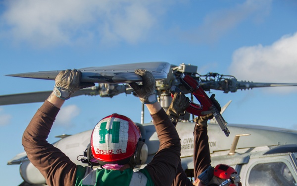 Two naval aviation deck crew members in green vests and red helmets are manually folding or unfolding a helicopter rotor blade on what appears to be an aircraft carrier flight deck. The helicopter visible in the background bears markings referencing CVW (Carrier Air Wing) and 'George Washington,' suggesting this is aboard USS George Washington (CVN-73). The crew members are lifting the rotor blade overhead in a coordinated effort.