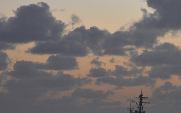 A naval warship is silhouetted against a dramatic twilight sky over open ocean. The vessel appears to be underway, photographed from a distance at either dawn or dusk with orange-tinted clouds on the horizon. The ship's profile suggests it is a modern destroyer or frigate-class vessel.