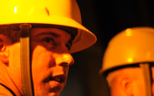 Two workers wearing yellow hard hats are photographed in a low-light environment with a strong warm orange/amber light source illuminating their faces from below or the side. Both individuals are wearing chin straps on their helmets and appear to be in an industrial or construction setting. The foreground subject is in sharp focus while the second worker is slightly blurred in the background.