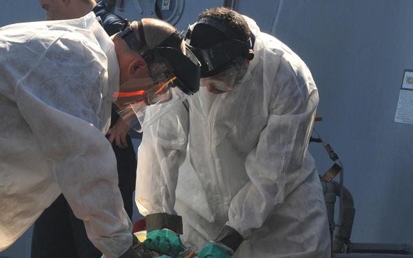 Two personnel wearing white Tyvek protective suits, respirators/gas masks, and safety goggles are examining a large cylindrical object secured on yellow cradle stands. The object appears to be a munition or ordnance item with red banding markings, resting on what appears to be a naval vessel deck. The scene suggests an ordnance inspection, EOD (Explosive Ordnance Disposal), or hazardous materials handling procedure.