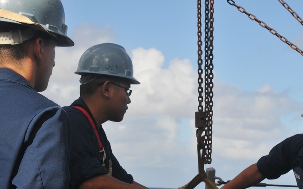 Three U.S. Navy personnel in blue coveralls and gray hard hats are handling a large aerial bomb or ordnance on the deck of a naval vessel at sea. The munition is suspended by a chain hoist/crane and is being guided into position by the sailors. The operation appears to be an ordnance transfer or handling evolution conducted aboard ship.