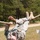 A U.S. Army soldier in ACU (Army Combat Uniform) is traversing a rope obstacle course, hanging upside down from a horizontal rope. The soldier is wearing tan combat boots and has a U.S. flag patch visible on the left sleeve. The setting appears to be an outdoor military training area with grass, a dirt path, and trees in the background.