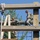 Two military personnel in US Army ACU (Army Combat Uniform) camouflage are navigating a wooden obstacle course structure. One soldier is climbing over a horizontal log obstacle while the other, wearing a patrol cap and safety harness, observes or assists. The structure includes rope nets, wooden beams, and climbing ropes consistent with a military training facility.