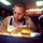 Life after formation: Veteran finds success in tattoo art A heavily tattooed individual is leaning over a work surface in what appears to be a tattoo parlor, focused on drawing or sketching a design. The person is wearing a white sleeveless shirt and has visible tattoos covering their arms, chest, and neck, along with facial piercings. A neon 'TATTOO' sign is illuminated in the background.