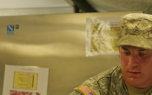 A U.S. Army soldier identified by the name tape 'YOUNG' is reviewing documents in what appears to be a military field kitchen or food service facility. He is wearing the Army Combat Uniform (ACU) with digital camouflage pattern and holds the rank of Sergeant, visible on his patrol cap. The background shows stainless steel equipment with posted notices and documents.