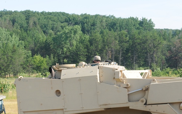 A sand/tan-colored armored combat engineering vehicle, consistent with an M1 Assault Breacher Vehicle (ABV) or similar combat engineer platform, is shown on a grassy field area towing a green military trailer. At least one soldier is visible in the open hatch of the vehicle. A second military truck with a canvas cover is visible in the background to the right.