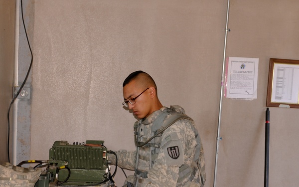 A U.S. Army soldier in ACU (Army Combat Uniform) is standing at a field desk operating what appears to be military radio communications equipment. The setting appears to be a forward operating base or field command post interior. The soldier is wearing glasses, a tactical vest, and has a combat helmet resting on the desk beside the equipment.
