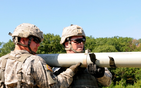 Two U.S. Army soldiers in ACU (Army Combat Uniform) digital camouflage are handling a cylindrical munition or missile canister, likely loading or unloading it from a military vehicle. Both soldiers are wearing Kevlar helmets, tactical vests, and protective eyewear. The setting appears to be a training range or field exercise area with green vegetation in the background.