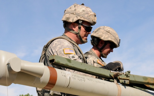 Two or more U.S. Army soldiers in ACU (Army Combat Uniform) pattern camouflage are handling a large missile or rocket system, likely loading or securing it onto a vehicle or platform. The soldiers wear American flag patches on their shoulders, combat helmets, and tactical gear. The setting appears to be an outdoor training area or range with green vegetation visible in the background.