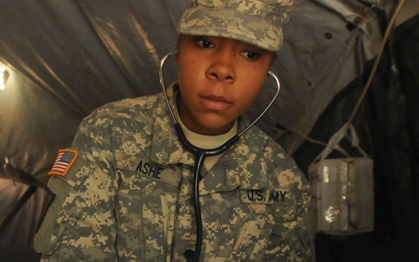 A U.S. Army soldier named ASHE, wearing ACU (Army Combat Uniform) digital camouflage and a stethoscope around their neck, is performing a medical procedure inside what appears to be a military field tent. The soldier is wearing purple medical gloves and attending to what appears to be a simulated casualty represented by a fur-covered mannequin or training dummy with white bandaging/dressing applied. This appears to be a medical training exercise.