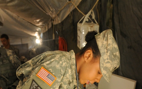 A U.S. Army medical personnel wearing ACU (Army Combat Uniform) with a stethoscope is working at a field medical station inside a military tent. The soldier is leaning over a table examining or treating what appears to be a simulated casualty (training mannequin or moulage) while surrounded by medical supplies. The setting appears to be a field medical training exercise or forward operating base aid station.