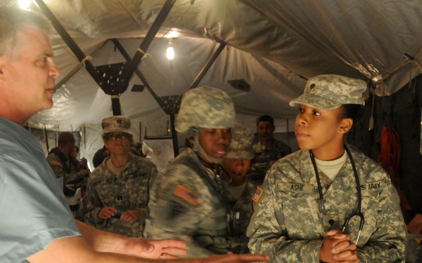 A medical training or briefing session inside a military field tent, where a civilian medical professional in scrubs appears to be instructing a group of U.S. Army soldiers. A soldier with a stethoscope and name tape reading 'ASHE' is prominently featured, suggesting a medical corps training exercise. A simulated casualty or training mannequin covered in fur-like material is visible on the table in the foreground.