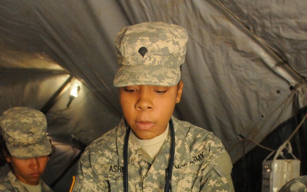 A U.S. Army soldier with the last name 'Ashe' wearing ACU (Army Combat Uniform) and a stethoscope administers an injection via syringe to a large canine animal, likely a dog or coyote, inside a military tent. A second soldier in the background holds a clipboard, apparently recording observations. The setting appears to be a field veterinary or medical training exercise.