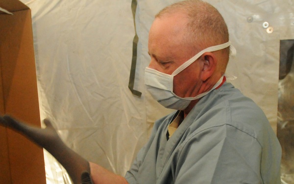 A male healthcare worker wearing blue scrubs and a surgical face mask is putting on or adjusting dark nitrile examination gloves. He appears to be in a temporary or field medical setting, indicated by the plastic sheeting visible in the background. Medical supplies and packaging are visible nearby.
