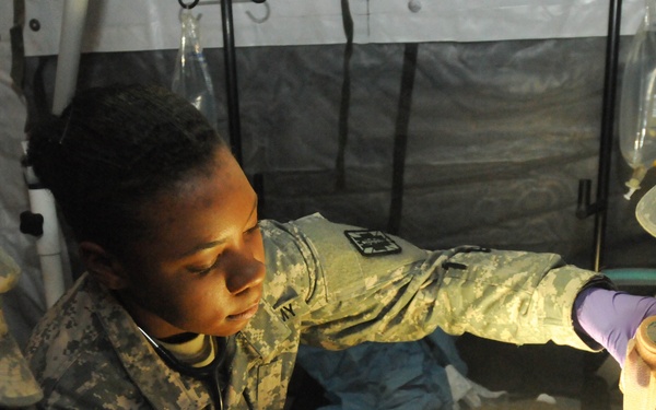 U.S. Army soldiers in ACU (Army Combat Uniform) camouflage are performing what appears to be a medical training exercise inside a military field tent or aid station. At least three personnel are visible, with one wearing purple medical gloves and holding bandaging material over a simulated patient or training mannequin illuminated by a bright light. The setting includes IV bags hanging overhead, consistent with a field medical treatment facility.