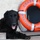 A black Labrador-type dog is lying down on a dock or platform in front of an orange life preserver ring. The life ring is labeled 'Coast Guard Station Honolulu,' indicating this photo was taken at a U.S. Coast Guard facility in Hawaii. The dog appears relaxed and happy with its tongue out, suggesting it may be a station mascot or working dog.