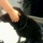 A wet black dog wearing a collar is standing on what appears to be the deck of a boat. A person's hand is resting on the dog's head, and another person's legs are visible in the lower right corner. The dog's fur is visibly damp, suggesting it has recently been in water.