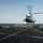 A white military helicopter is hovering just above a naval ship's flight deck, appearing to be in the process of landing or taking off. A flight deck crew member wearing a yellow safety vest and white helmet is visible on the left, directing the aircraft with an outstretched arm. The scene takes place over open ocean under clear skies.