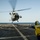 A helicopter is hovering over the flight deck of a U.S. Navy destroyer, designated DDG-87, during a landing or departure operation. A flight deck crew member wearing a yellow safety vest marked 'DDG 87' and a white helmet with hearing protection is signaling the aircraft. The scene takes place at sea under clear skies with calm ocean visible in the background.