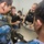 Military personnel from two different nations are gathered around a table in what appears to be a briefing or training session. A soldier in desert camouflage is holding and demonstrating an assault rifle (appears to be an M4/M16-style weapon) to personnel wearing blue digital camouflage uniforms bearing Chinese People's Liberation Army Navy (PLAN) insignia. This appears to be a joint military training or exchange exercise.
