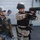 Two armed military personnel are visible in the foreground on what appears to be a ship's deck. One individual wearing blue digital camouflage and a blue helmet is standing while the other, in desert camouflage with a black helmet, is demonstrating a pistol aiming stance. A third person in blue camouflage is visible in the background.