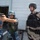The image shows two armed military personnel in full tactical gear on what appears to be the deck of a naval vessel. One soldier wearing blue digital camouflage and a blue helmet is aiming a pistol in a training or demonstration stance, while a second soldier in desert camouflage with a black tactical vest and black helmet observes. A third person in blue camouflage uniform is visible in the background near a large door or hatch.