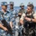 The image shows a military interaction between personnel wearing People's Liberation Army Navy (PLAN) uniforms and a Western military operator in tactical gear, taking place on the deck of a naval vessel at sea. The Western operator appears to be demonstrating or explaining equipment, possibly tactical vest components, to the Chinese naval personnel. This appears to be a joint military exercise or exchange event.