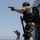 Military personnel are conducting a live-fire or training exercise on the deck of a naval vessel at sea. The foreground figure is heavily equipped in desert camouflage and tactical gear, aiming a pistol, while a second figure in blue camouflage aims a rifle in the background. Observers and a photographer are visible near a ship's superstructure on the right side.