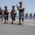 Four heavily equipped military personnel in desert camouflage uniforms and black tactical gear stand in a line on what appears to be a naval vessel's flight deck or open deck area. They are armed and wearing ballistic helmets, knee pads, and tactical vests. In the background, four additional personnel in blue/gray uniforms observe from the perimeter railing.