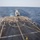 A white helicopter is landing or hovering just above the flight deck of a U.S. Navy destroyer at sea. Two deck crew members wearing helmets and colored vests are visible on the flight deck, with one signaling to the helicopter. The ship's hull number 'DDG 67' is partially visible on one crew member's vest.