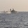 A naval warship is underway at speed on open ocean, generating a visible white wake and spray. A military helicopter is flying low alongside or near the vessel, possibly conducting a training exercise or operational maneuver. The scene appears to be a naval operation in calm but hazy open-sea conditions.