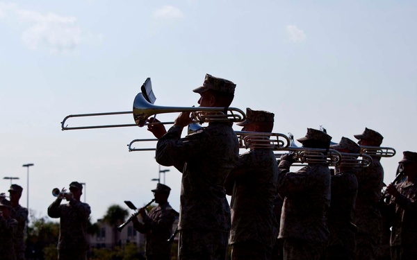 A military band is performing outdoors on a grass field, with personnel dressed in camouflage uniforms and caps. The musicians are playing brass instruments including trombones, trumpets, and baritone/euphonium horns. The scene appears to be a formal military ceremony or rehearsal at a military installation.