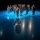 Lt. Gen. Dana Kyle Chipman retirement ceremony A military marching band is performing under dramatic blue stage lighting on a reflective floor. The ensemble is arranged in a semicircular formation with a drum major at the front wearing a distinctive tall black bearskin hat. The scene is captured from a low angle, emphasizing the drum major's shadow stretching toward the camera.