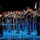 Lt. Gen. Dana Kyle Chipman retirement ceremony A military ceremonial band performs on a stage illuminated by blue lighting. Musicians in formal dress uniforms with red-crowned caps and gold trim play brass instruments including trombones, euphoniums, and tubas. A drum major stands at the left holding a ceremonial mace, while color guard members with flags are visible in the background.