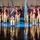Lt. Gen. Dana Kyle Chipman retirement ceremony A formal military ceremony featuring soldiers dressed in 18th-century Continental Army-style uniforms with tricorn hats, white crossbelts, and red-trimmed blue coats. The soldiers are arranged in formation on a highly reflective floor that mirrors their figures below. A light blue flag with stars is prominently displayed at center, and multiple colored flags line the background wall.