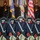 Lt. Gen. Dana Kyle Chipman retirement ceremony Five soldiers dressed in 18th-century Continental Army ceremonial uniforms stand in formation during a military color guard ceremony. They are viewed from behind, carrying muskets and wearing tricorn hats, white crossbelts, and red haversacks. In the background, modern military personnel in contemporary dress uniforms are visible alongside multiple flags including the American flag.