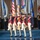 Lt. Gen. Dana Kyle Chipman retirement ceremony A formal military color guard ceremony is taking place indoors, featuring soldiers dressed in 18th-century Continental Army-style uniforms marching in formation while carrying flags and rifles. Behind them, additional military personnel in modern dress uniforms stand at attention flanking a large display of state and organizational flags. The event appears to be a formal military ceremony or retreat held in a large convention or ceremonial hall.