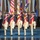 Lt. Gen. Dana Kyle Chipman retirement ceremony A formal military color guard ceremony featuring soldiers dressed in 18th-century Continental Army-style uniforms marching in synchronized formation. The foreground soldiers wear distinctive tricorn hats, red and navy blue coats with white cross-belts, and carry muskets with fixed bayonets. Behind them, additional military personnel in modern dress uniforms stand at attention holding a large array of flags including the U.S. flag, state flags, and military branch flags.