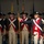 Lt. Gen. Dana Kyle Chipman retirement ceremony The image depicts a formal military reenactment or ceremonial drill performance featuring soldiers dressed in 18th-century American Revolutionary War-era uniforms. A central figure, distinguished by a navy blue coat with red facings, epaulettes, and white breeches, holds a ceremonial pike or spontoon, suggesting an officer's role. Rows of uniformed soldiers in red coats with white cross-belts and tricorn hats stand in formation behind him.