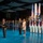 Lt. Gen. Dana Kyle Chipman retirement ceremony A formal U.S. military ceremony is taking place in a large indoor venue with a reflective blue floor. The scene includes senior military officers in dress uniforms, a civilian woman, and a color guard unit dressed in historical Revolutionary War-era uniforms carrying the U.S. flag and military unit colors. Rows of uniformed soldiers stand at attention in the background.