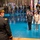 Lt. Gen. Dana Kyle Chipman retirement ceremony A formal U.S. Army military ceremony is taking place in a large indoor venue with a distinctive blue floor. A senior officer in dress uniform is facing two individuals — a uniformed general and a civilian woman — who are standing at attention on the floor. The background features rows of soldiers in dress uniforms holding numerous state and military flags.
